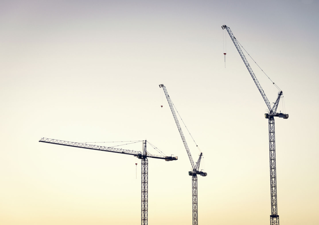 Three large construction cranes at a large industrial building site, photographed at dawn.