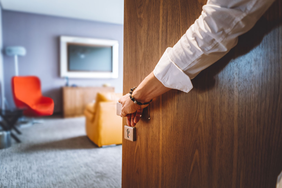 Toned man closing the door of the luxurious hotel room, man wearing a bracelet and a white shirt, closing the wooden door, nice and luxurious hotel room in the background.